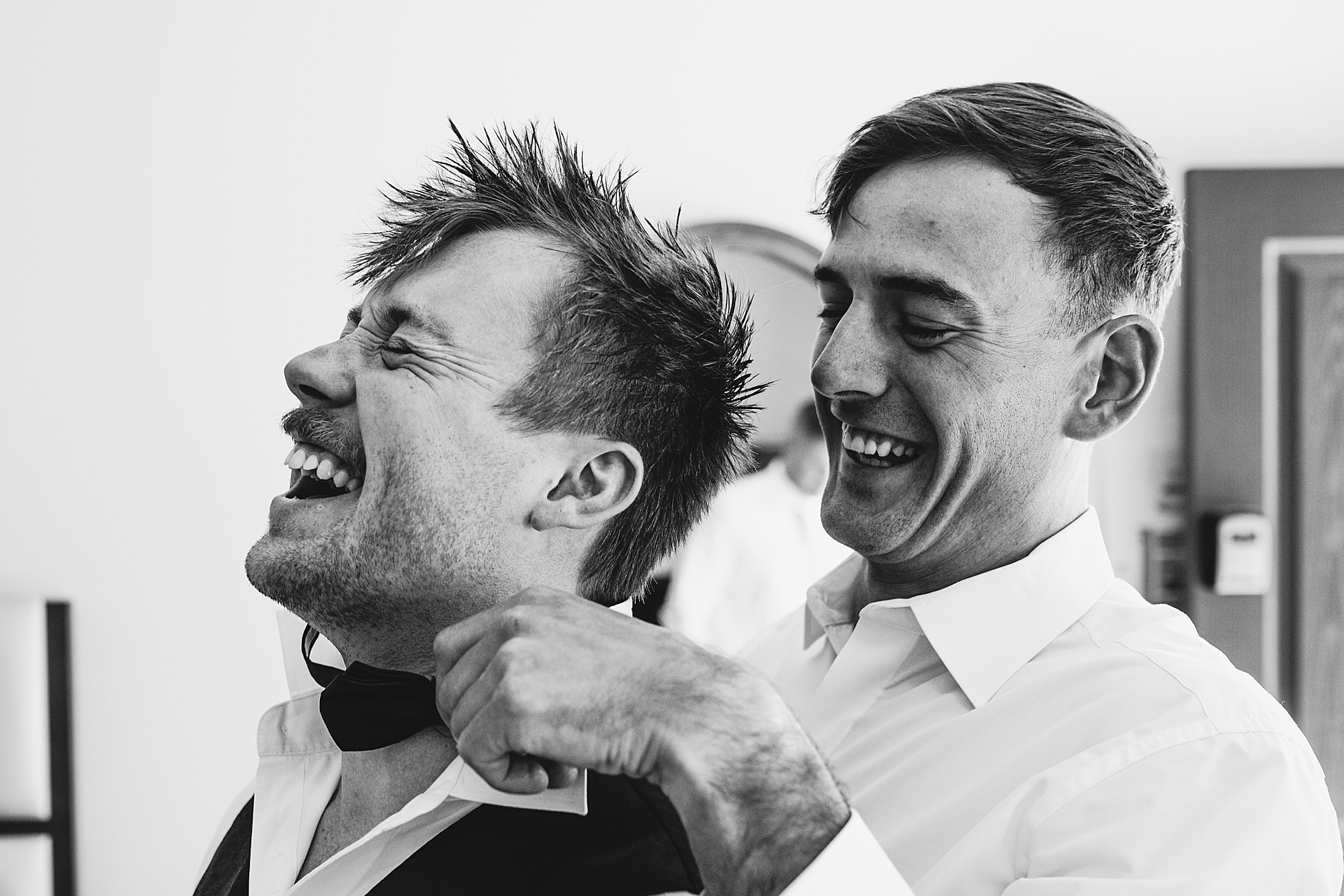 Black and white photo of a groomsman helping the groom with his bowtie while the groom laughs with his eyes closed. 