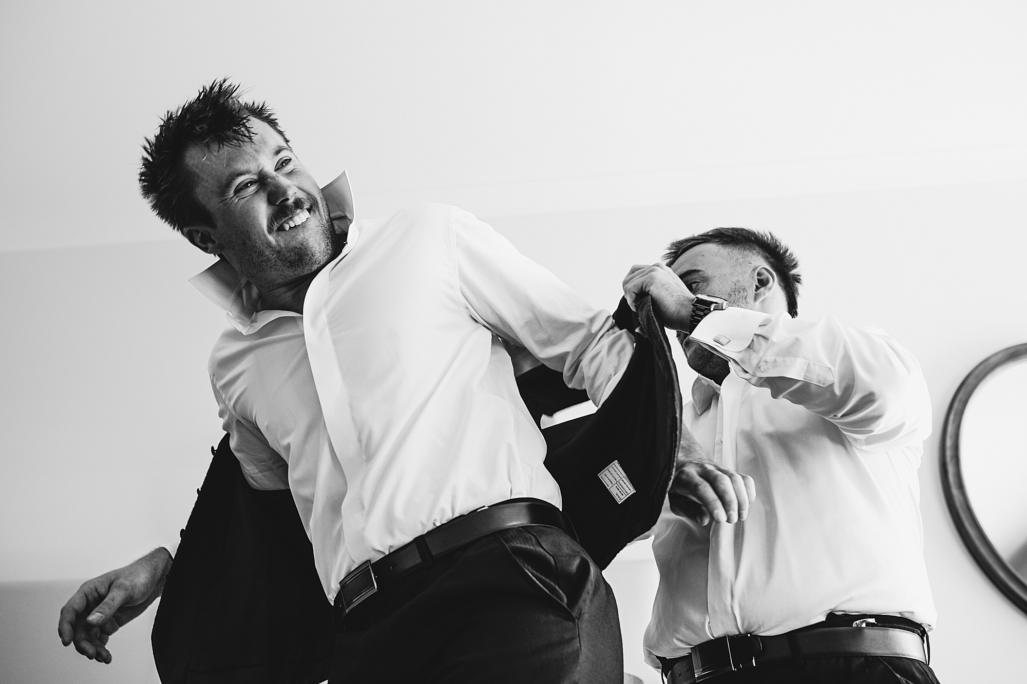 Black and white photo of a groom putting on his blazer and making a funny face while getting ready on his wedding day.