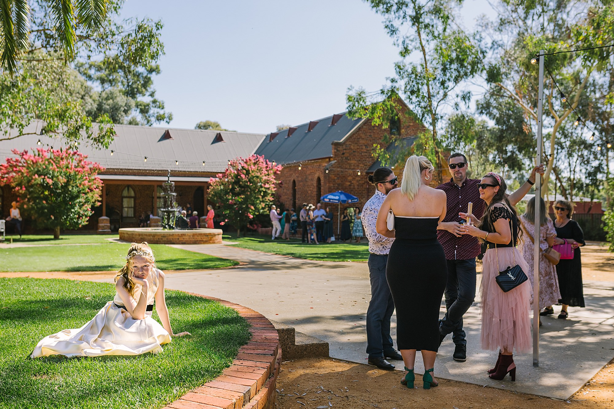 Young flower girl sitting on the grass while wedding guests enjoy drinks during cocktail hour.