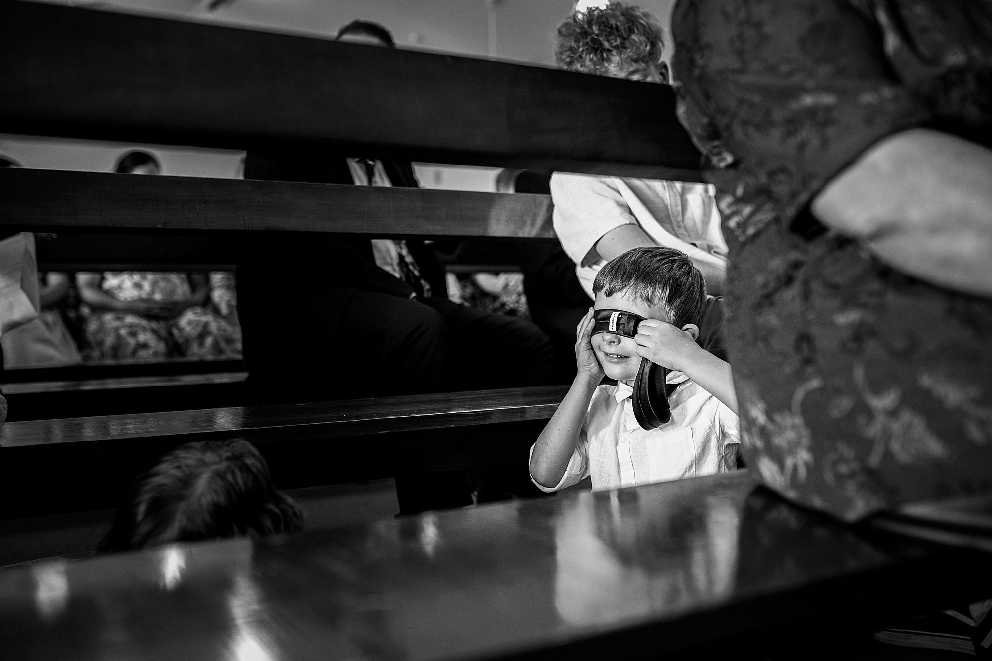 Black and white photo of a young child playfully holding his bow tie across his eyes during the wedding ceremony, while the bride and groom say their vows in the church.