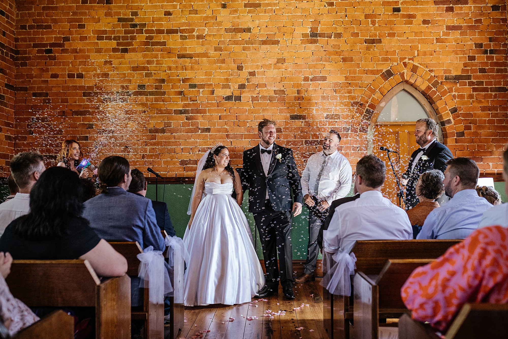 Bride and groom in a red brick chapel at Waratah Weddings, surrounded by bubbles as guests celebrate their just-married moment.