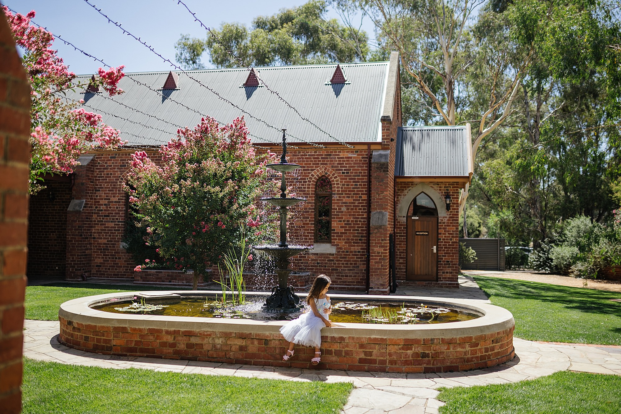 Little girl sitting by a pond in a garden with lush greenery and a red brick building in the background