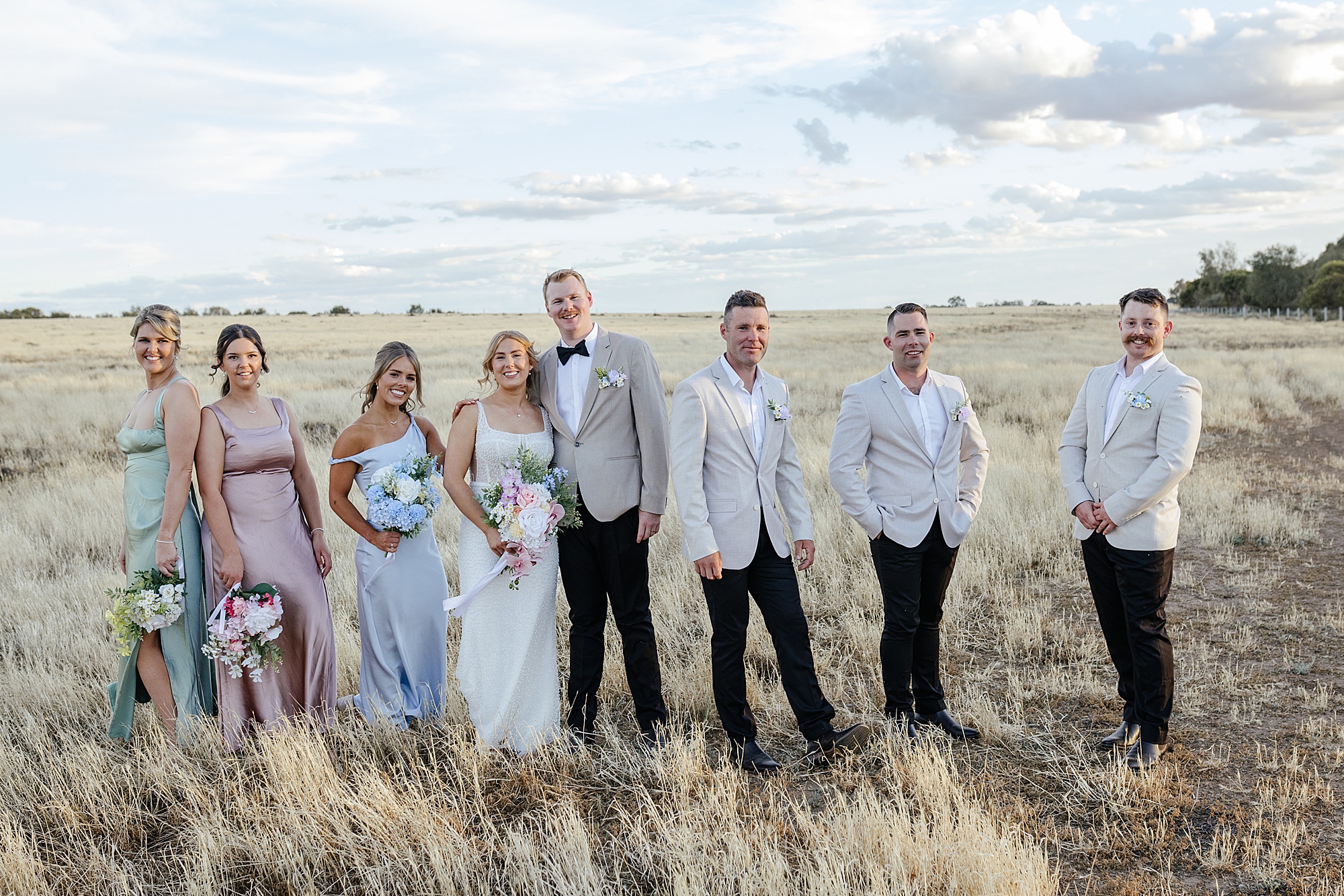 Large pastel coloured bridal party standing in open field during sunset.