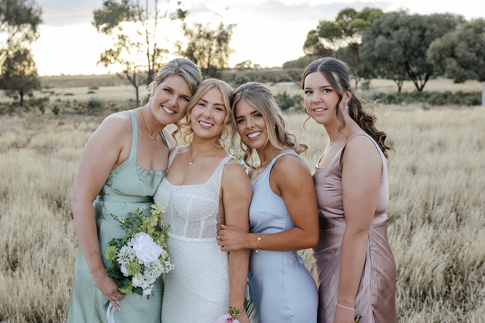 Bridesmaids in pastel dresses standing close to each other in open field.