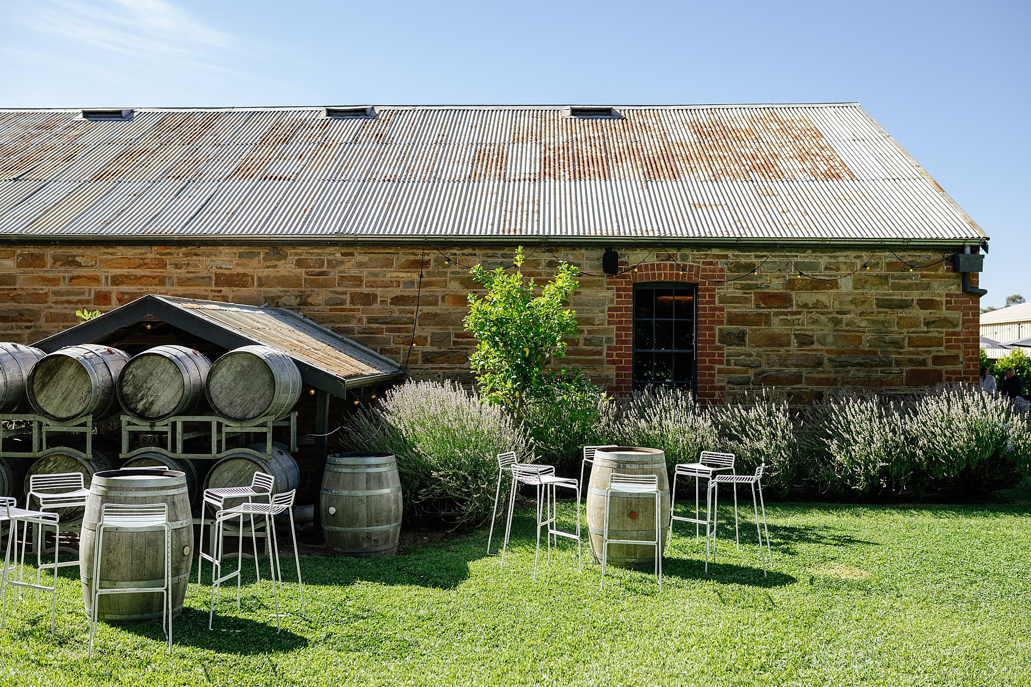 Sideview of sandstone building with barrels in a garden. 